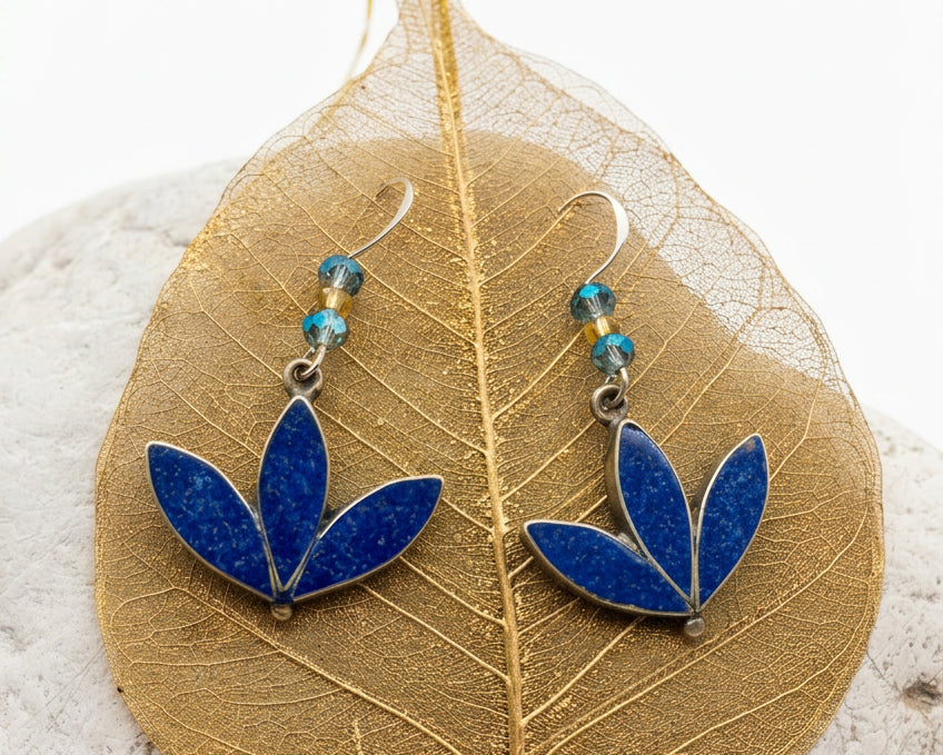 Blue enamel leaf-shaped dangle earrings with small blue and gold beads, displayed on a gold leaf over a white stone.