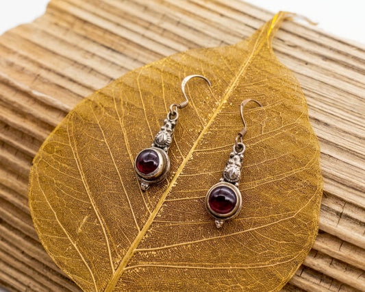 A pair of silver-toned dangle earrings with dark oval beads and small clear stones, displayed on a dried golden leaf over a woven straw mat against a white background.