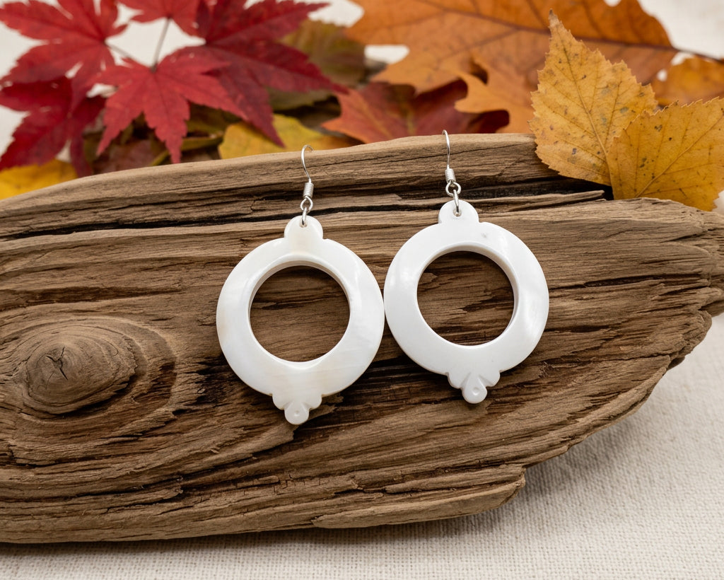 A pair of white circular drop earrings with silver fishhook wires, displayed on a piece of natural wood with red autumn leaves against a light gray background.