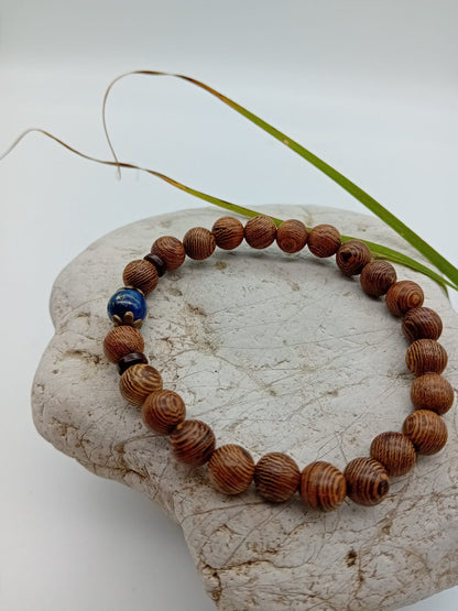 A beaded bracelet made of wenge brown wood beads with a single dark blue lapis lazuli bead and two small dark spacer beads, displayed on a light stone against a white background.