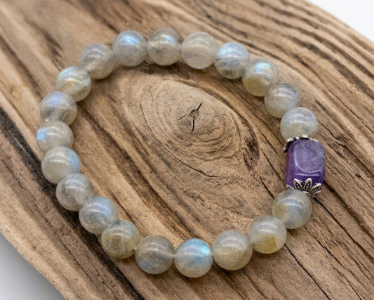 A beaded stretch bracelet made of round light gray-green stone beads with a prominent purple rectangular accent bead, displayed on a piece of natural wood against a white background.