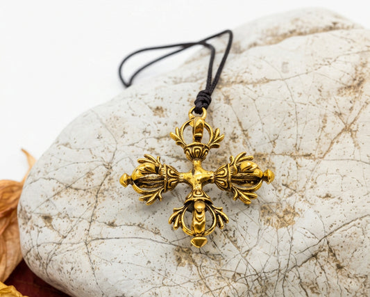 An ornate gold cross pendant on a black cord, displayed on a textured light gray stone with dried red and orange leaves on a white background.